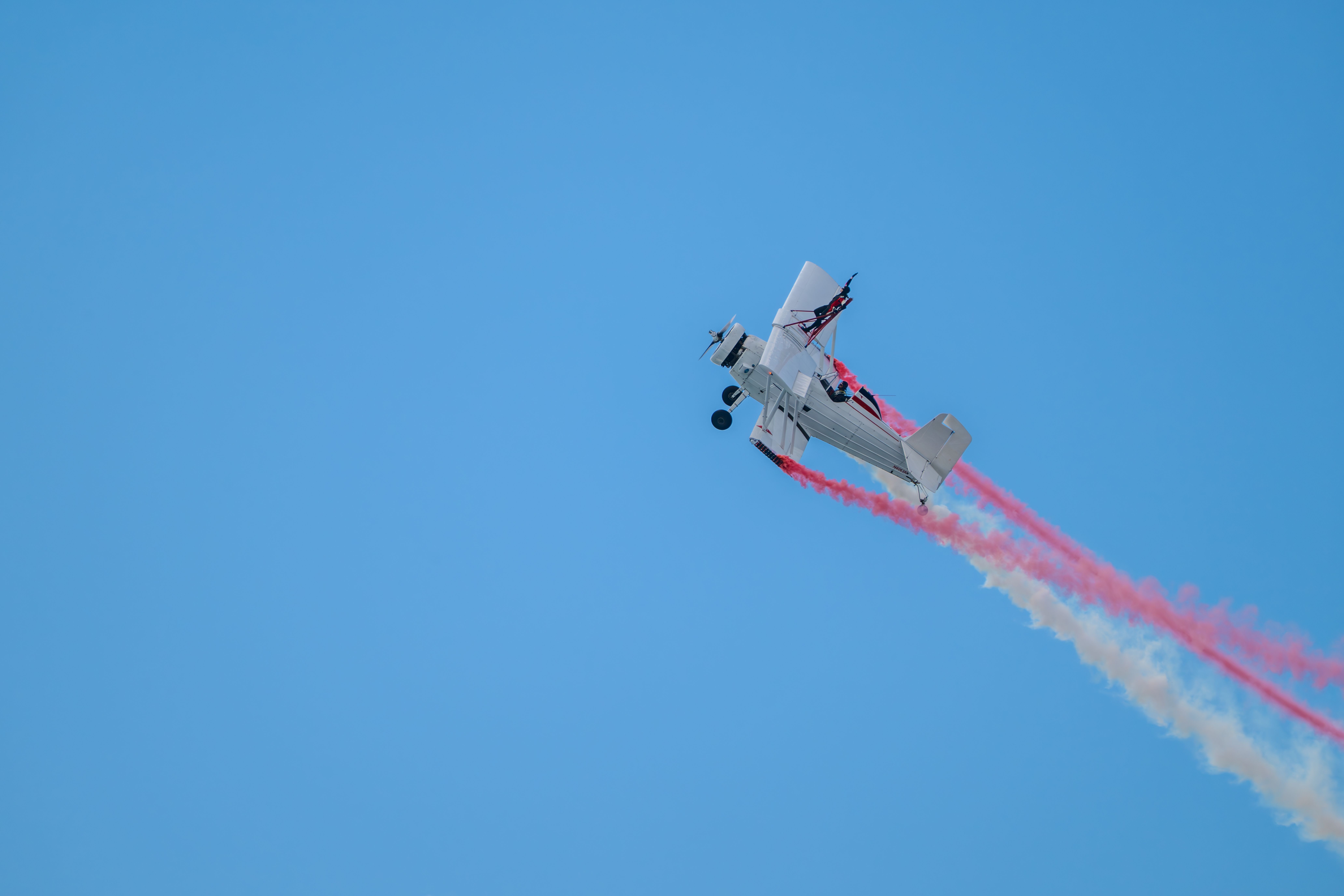 A vintage biplane performs an aerial stunt with colorful smoke trails against a clear blue sky.