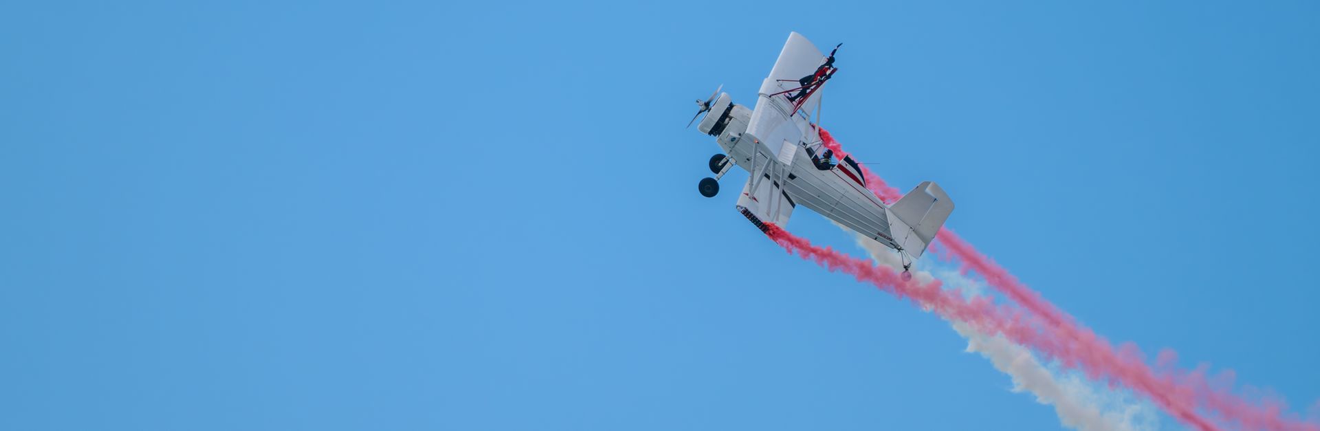 A vintage biplane performs an aerial stunt with colorful smoke trails against a clear blue sky.