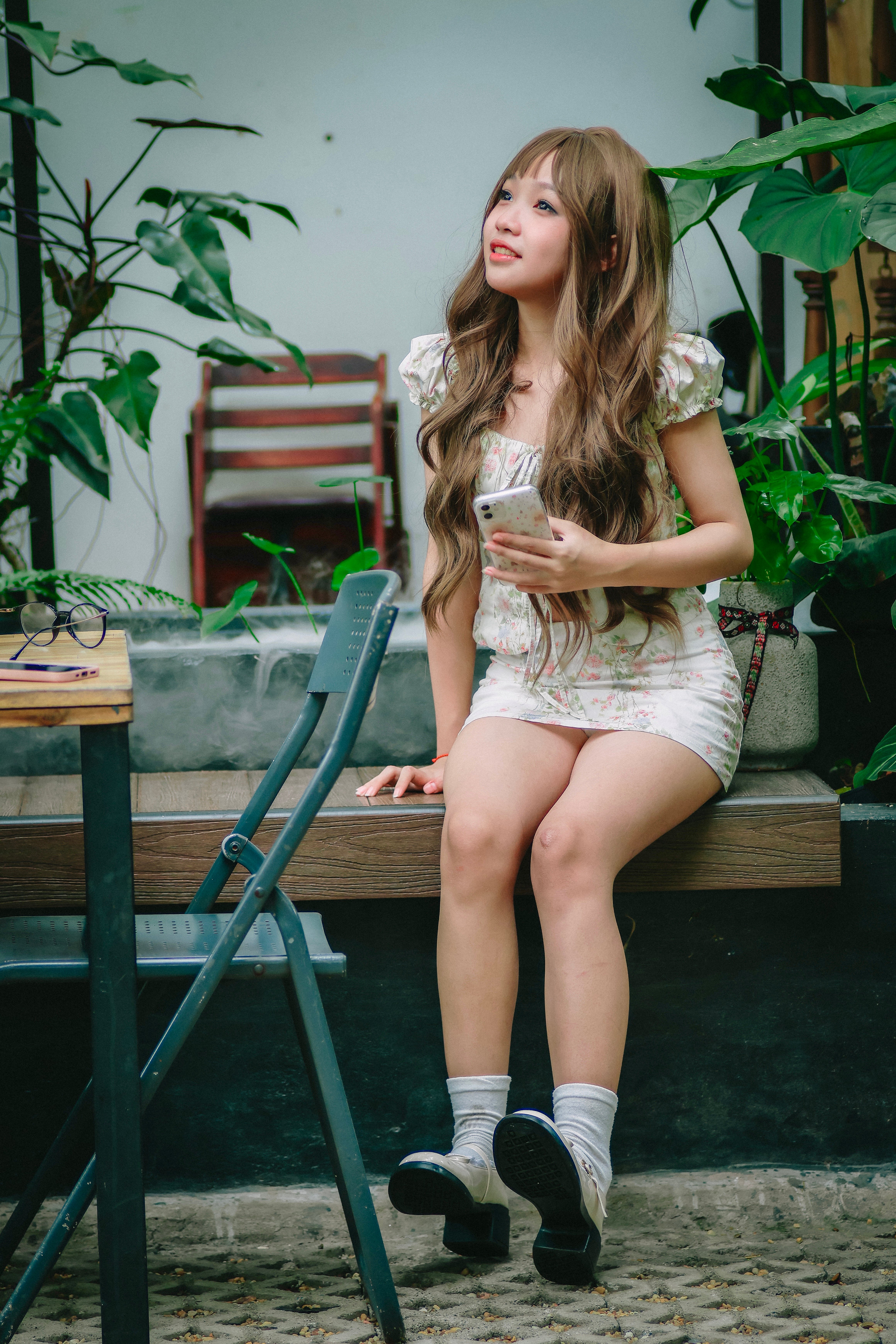 A young woman in a floral dress sits holding a smartphone at a chic outdoor cafe surrounded by lush greenery.