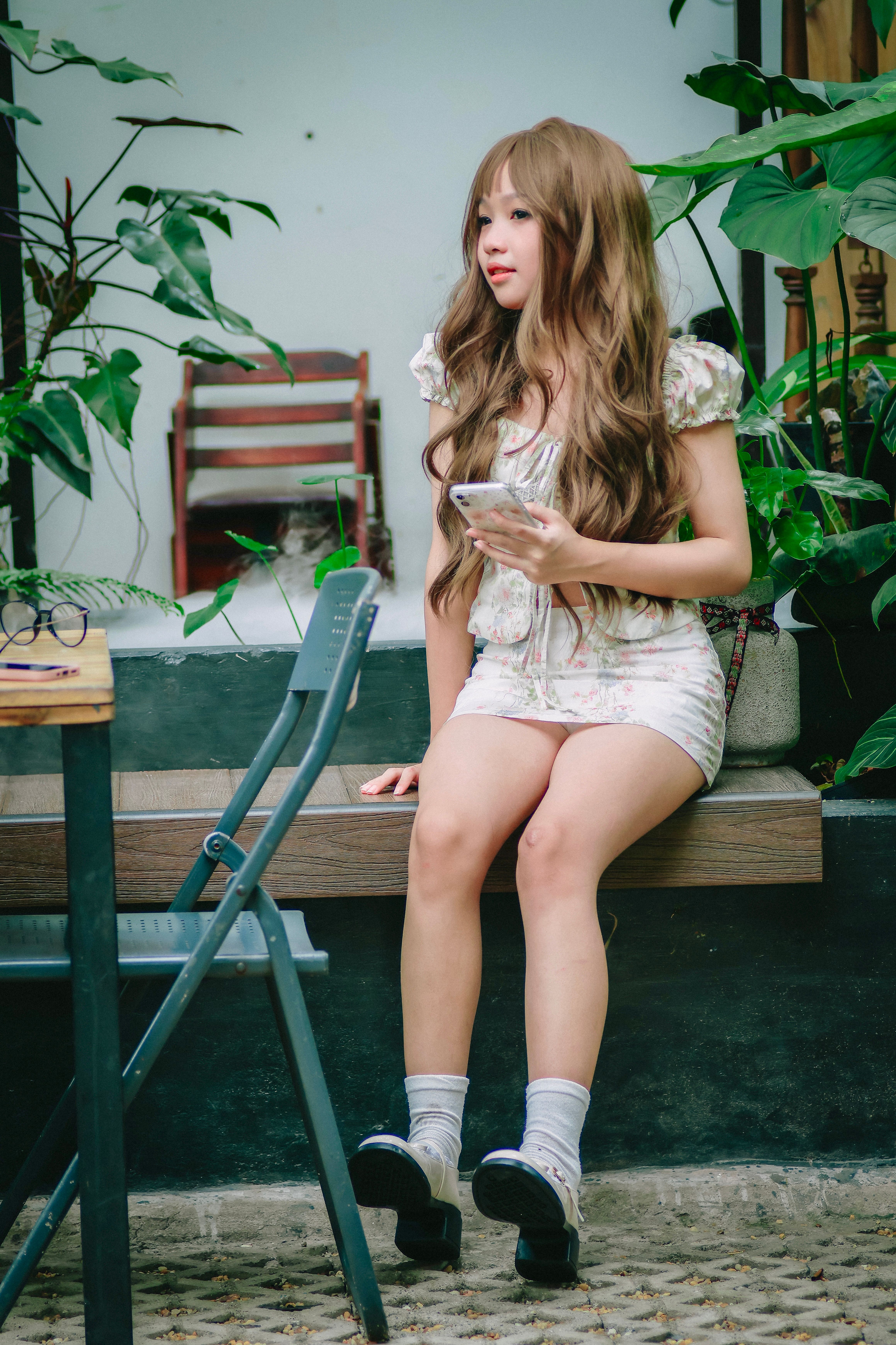 A young woman sitting on a bench holding a smartphone surrounded by lush greenery.