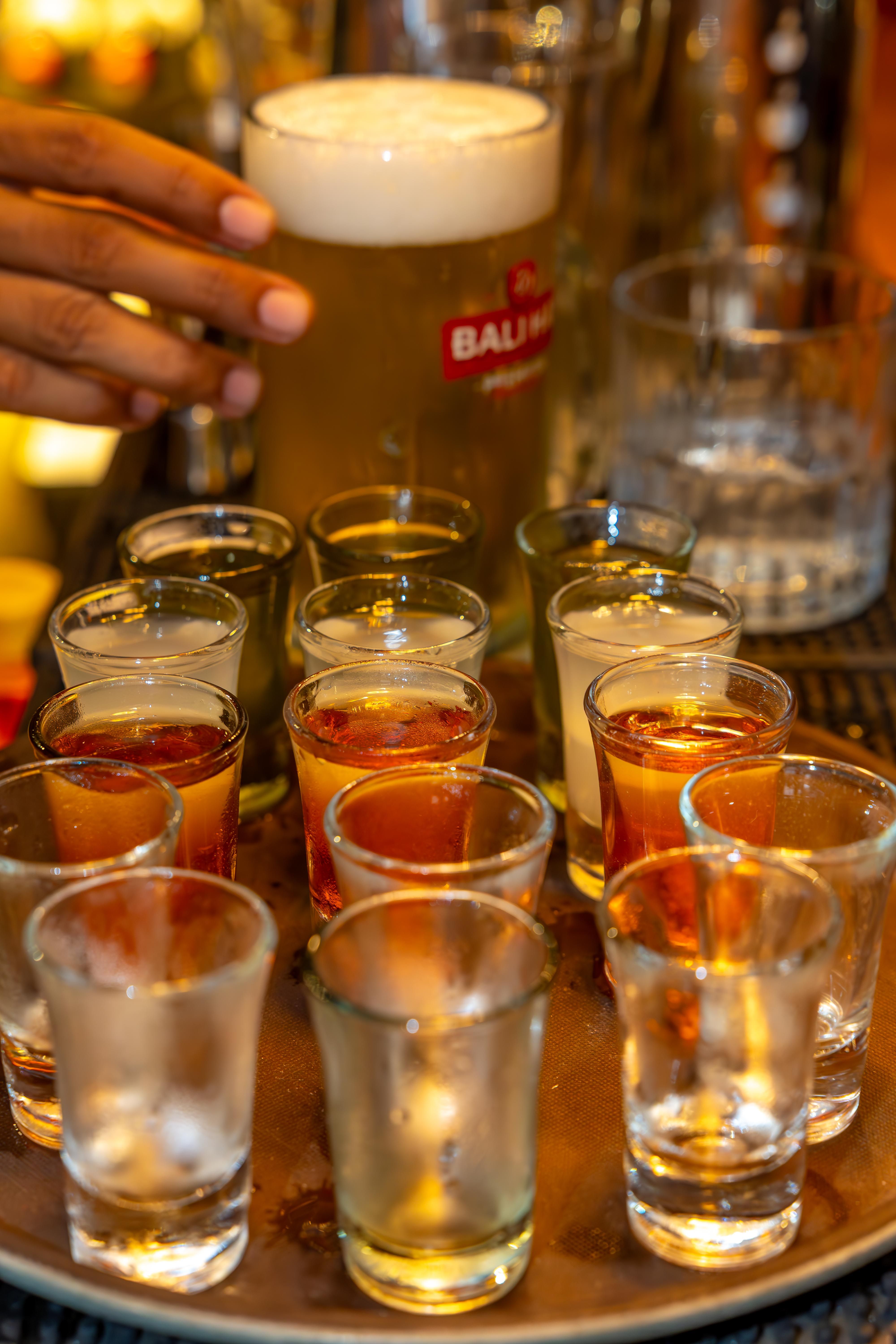 A hand reaches for a tall beer glass among various shot glasses on a bar tray.