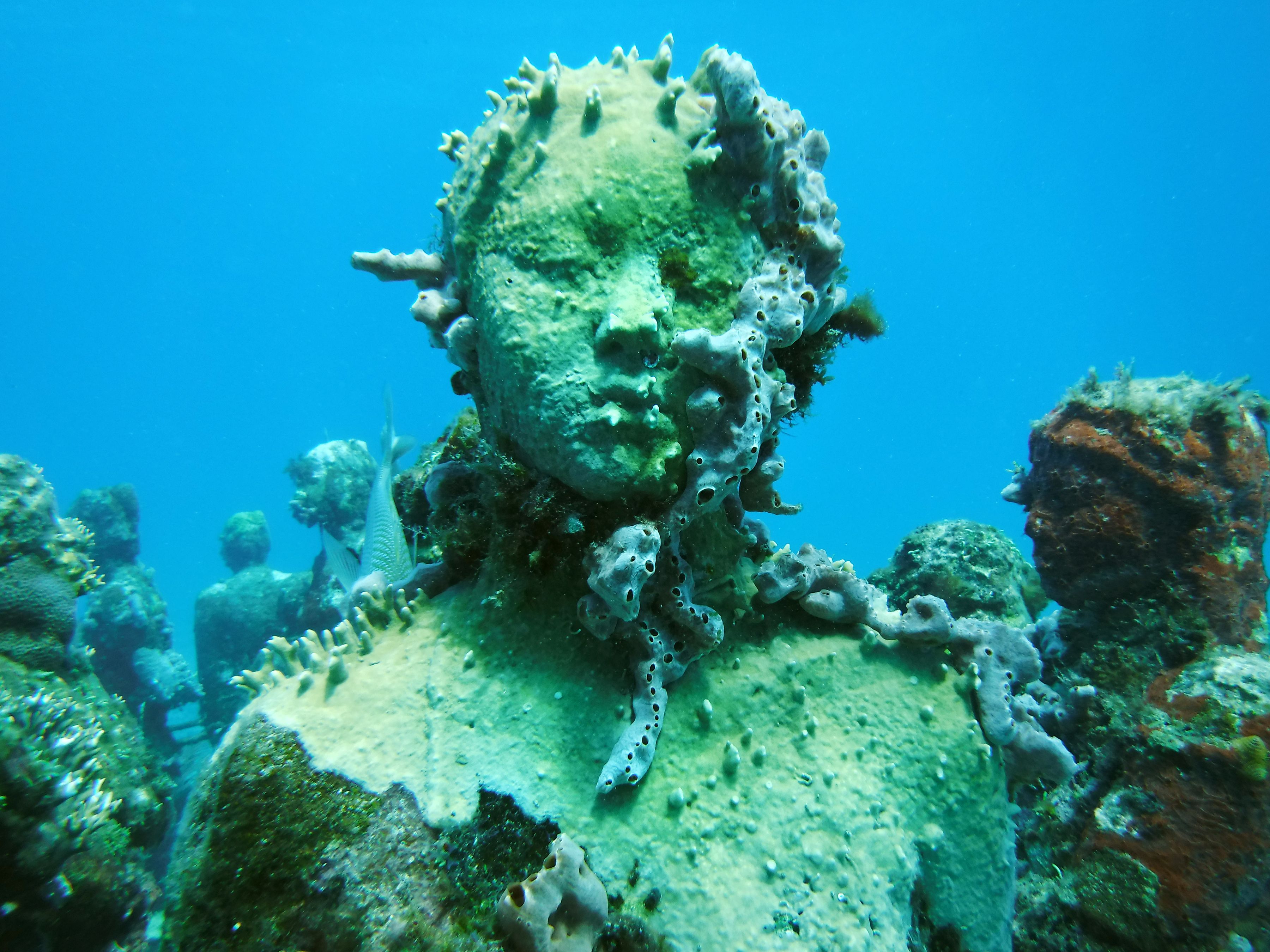 Marine-encrusted sculpture at Cancun Underwater Museum in Cancún, Mexico, shown underwater in clear blue sea.