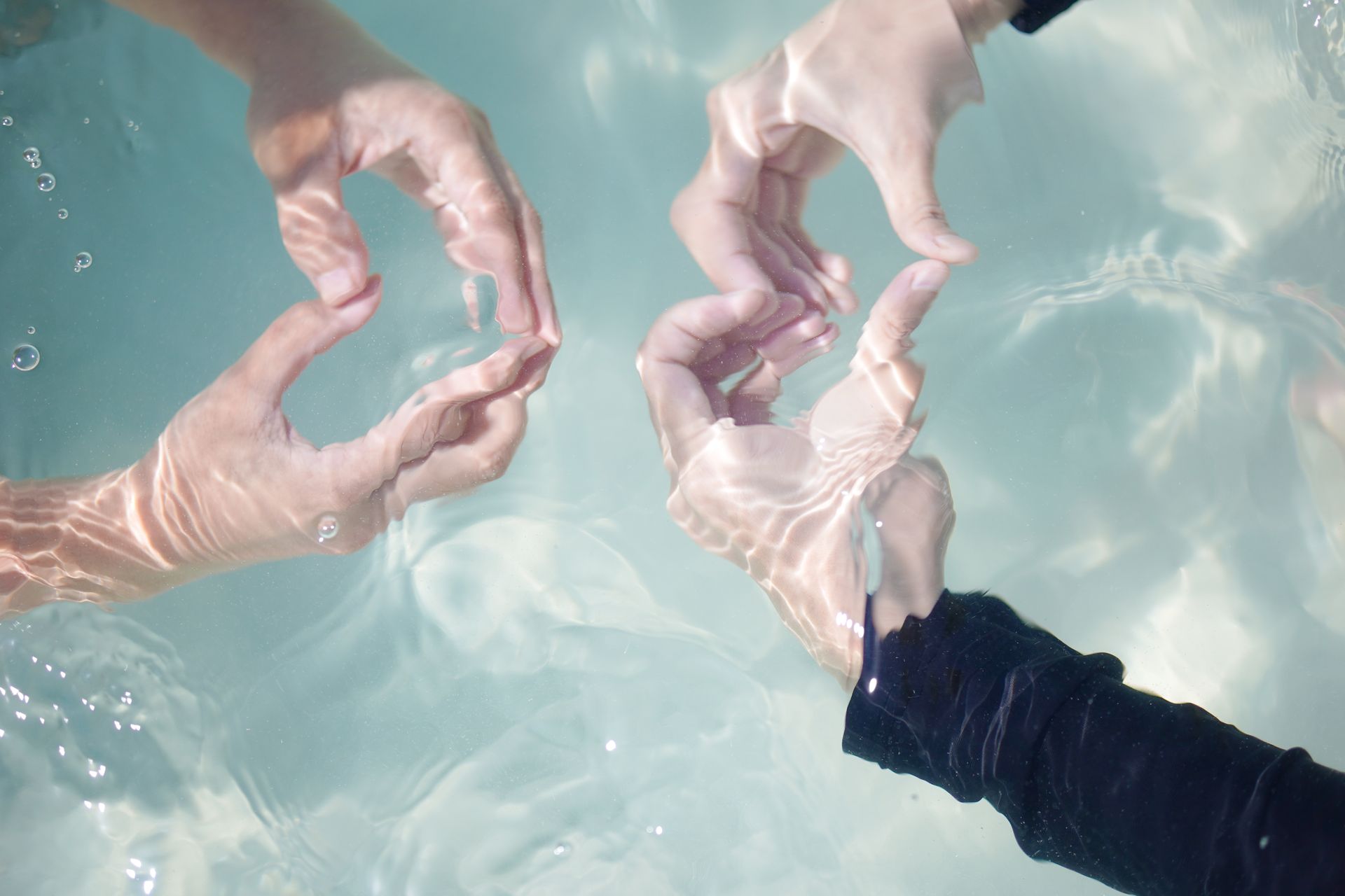 Three hands form shapes underwater with sunlight dancing on the skin.
