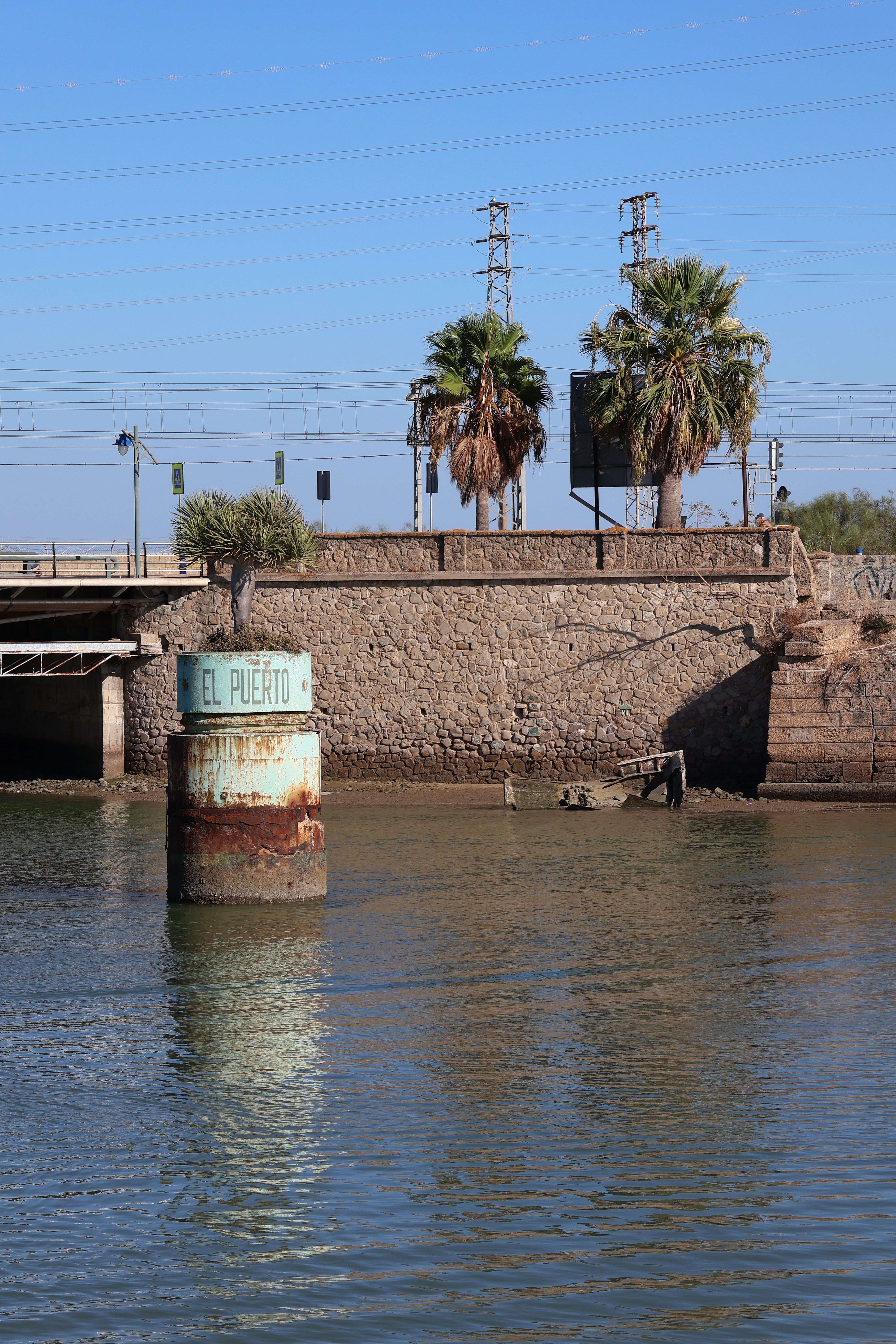 Riverside view in El Puerto de Santa María, showcasing palm trees and a stone wall.