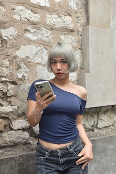 Stylish young woman with short hair holding a smartphone against a rustic stone wall.