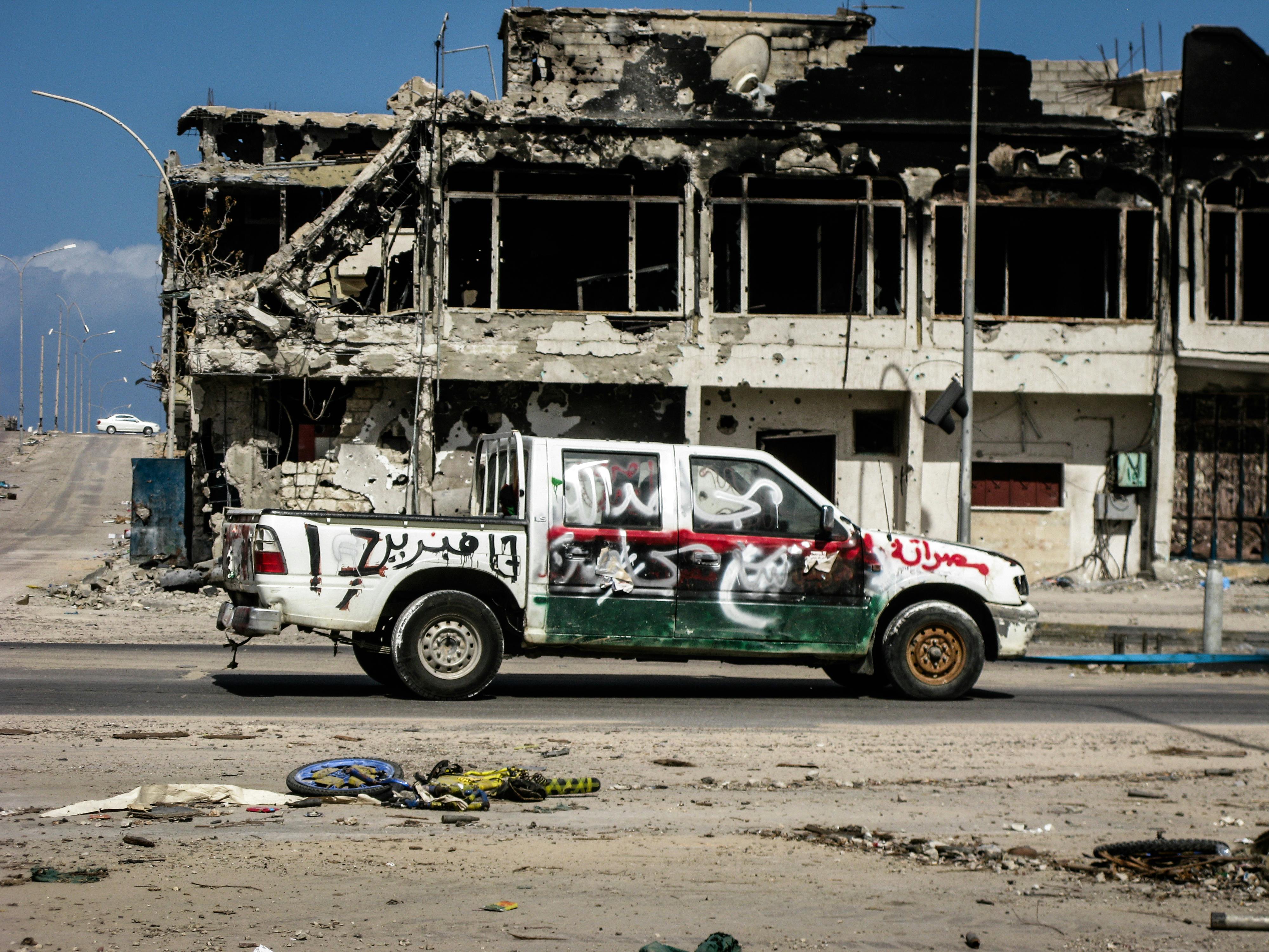 A damaged vehicle in front of a war-torn building in Sirte, Libya, depicting the impact of conflict.