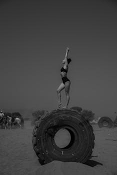 Woman in athletic pose on a giant tire in a desert landscape captured in black and white.