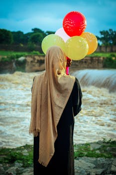 A woman in traditional attire holds balloons beside a river under a cloudy sky, evoking a serene and introspective mood.