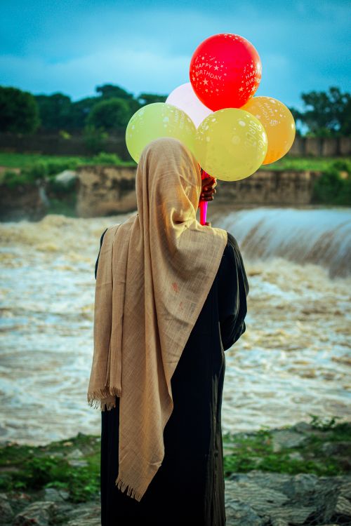 Free A woman in traditional attire holds balloons beside a river under a cloudy sky, evoking a serene and introspective mood. Stock Photo