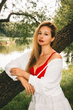 Portrait of a woman in a red dress leaning against a tree by a serene lake.