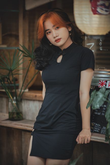 Stylish woman in black dress posing at a cozy cafe with plants and soft lighting.