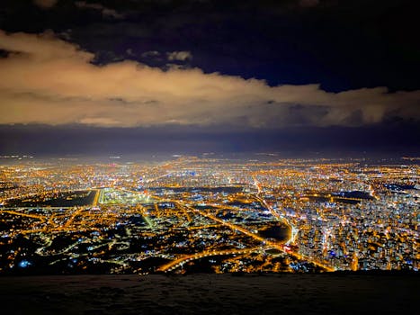 Aerial night view of a glowing cityscape with bright lights and cloudy skies.