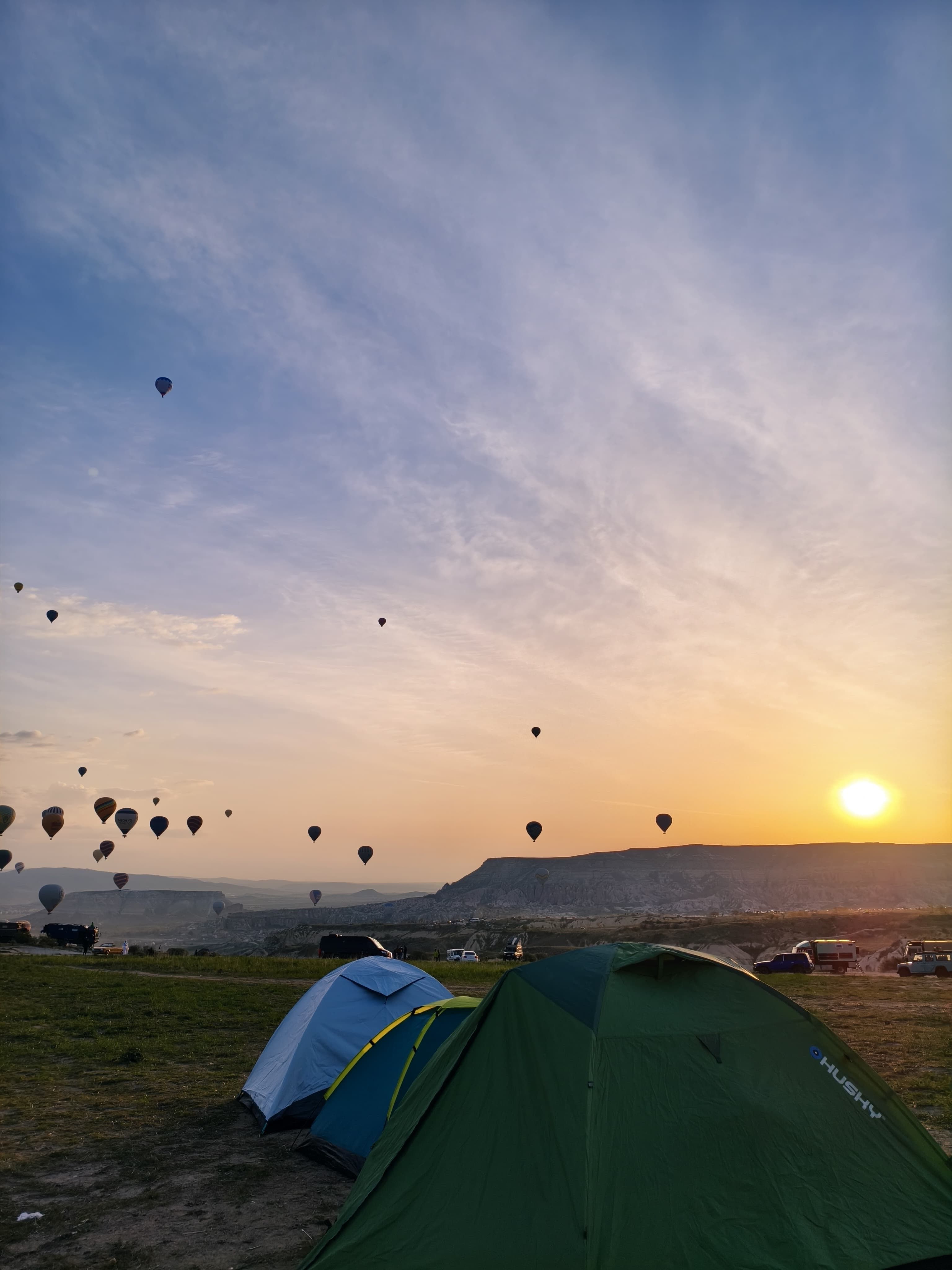 Peaceful sunrise with hot air balloons over tents in a serene camping area.