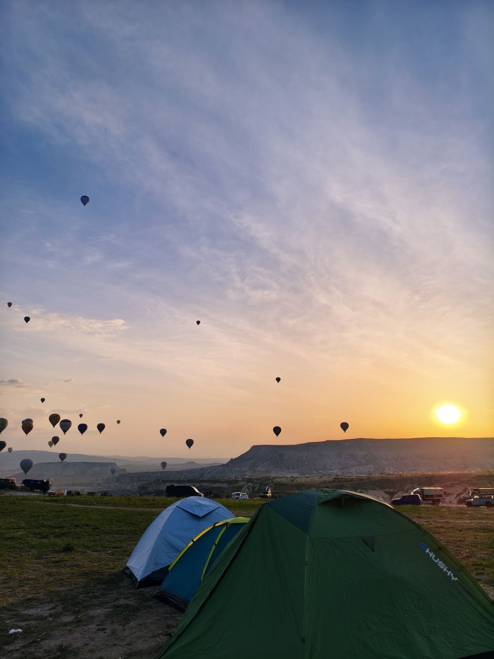 Peaceful sunrise with hot air balloons over tents in a serene camping area.