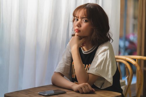 Free A young woman thoughtfully sitting at a wooden table near a window. Stock Photo