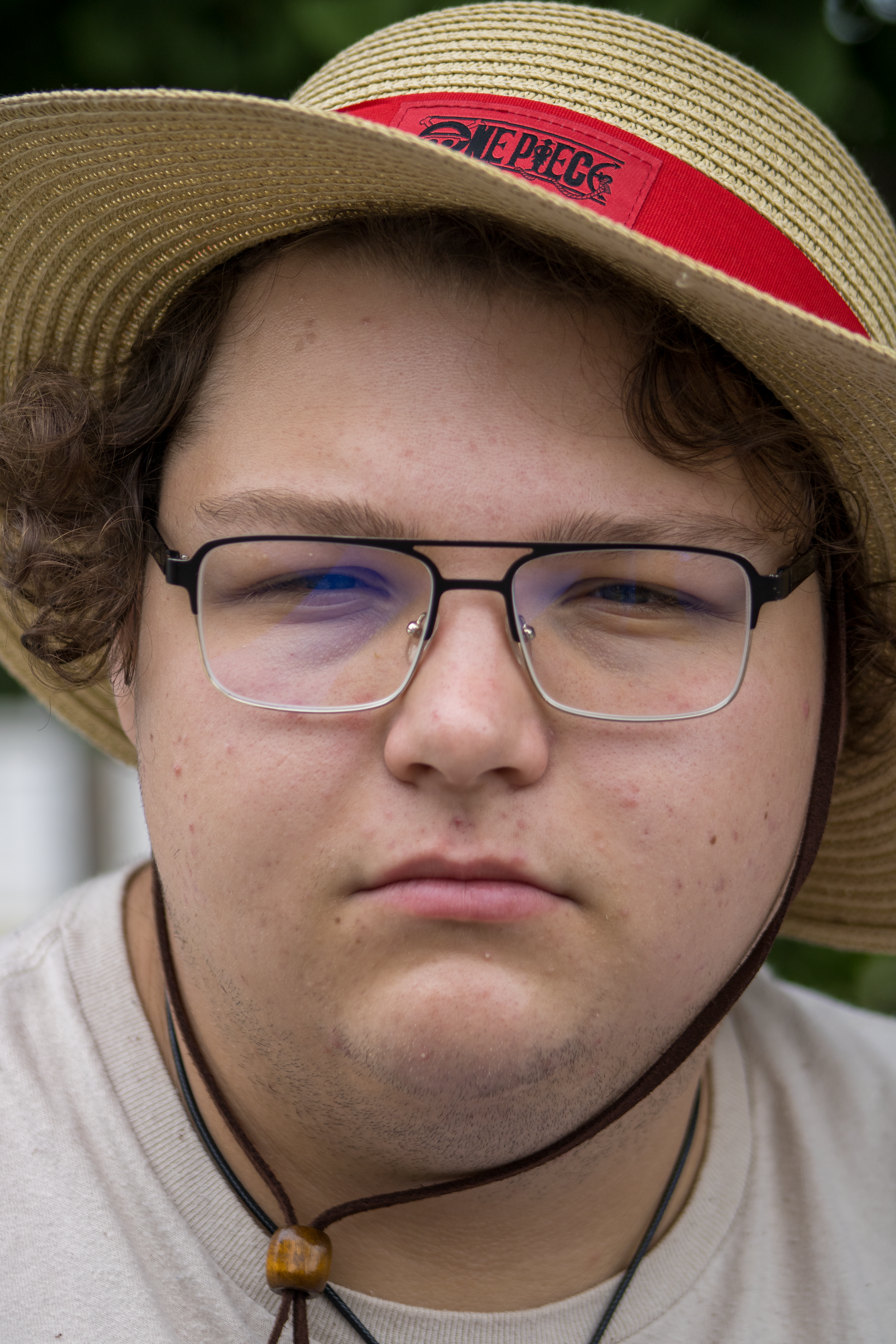 Close-up portrait of a young adult with glasses wearing a straw hat outdoors.