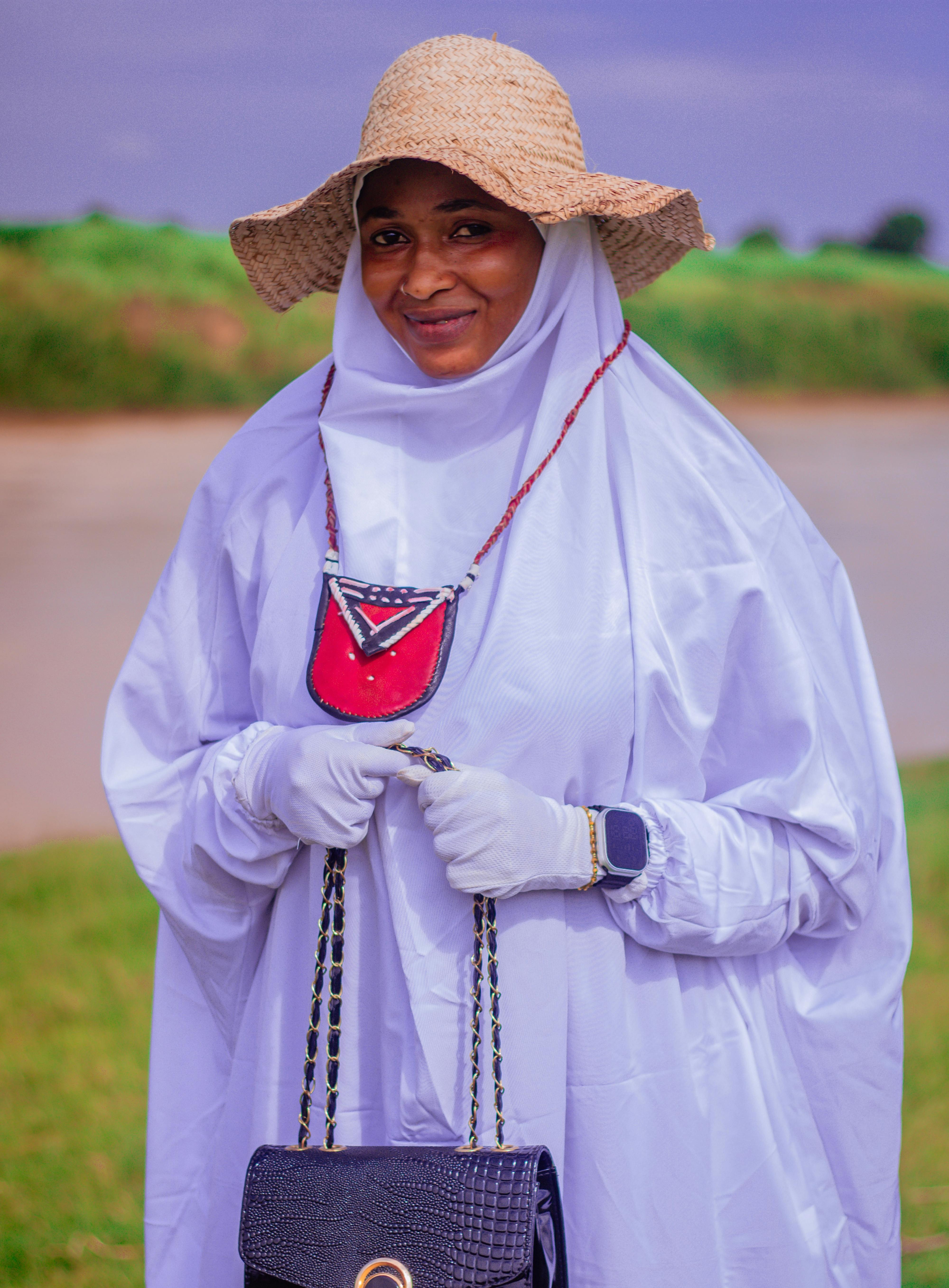 Smiling woman in white hijab, sun hat, and jewelry outdoors by the water.