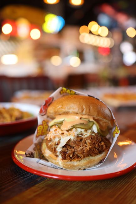 A glass jar of glossy yellow-orange Carolina Gold BBQ sauce with a spoon resting in it, next to a plate of pulled pork ready for basting.