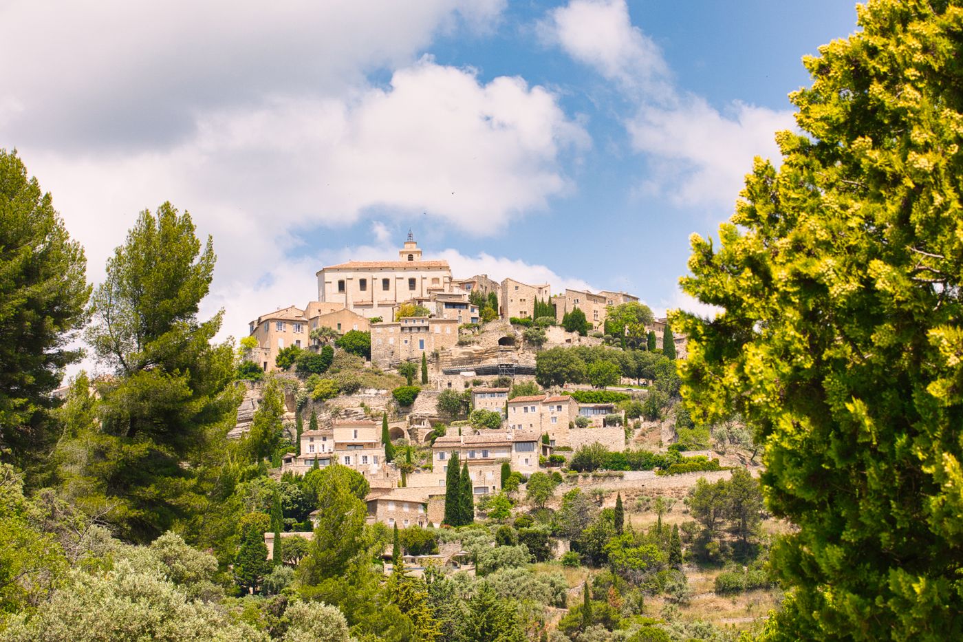 The hilltop village of Gordes in Provence, France - stone houses clustered densely on a hillside amid lush greenery, under a cloudy sky. No people visible.