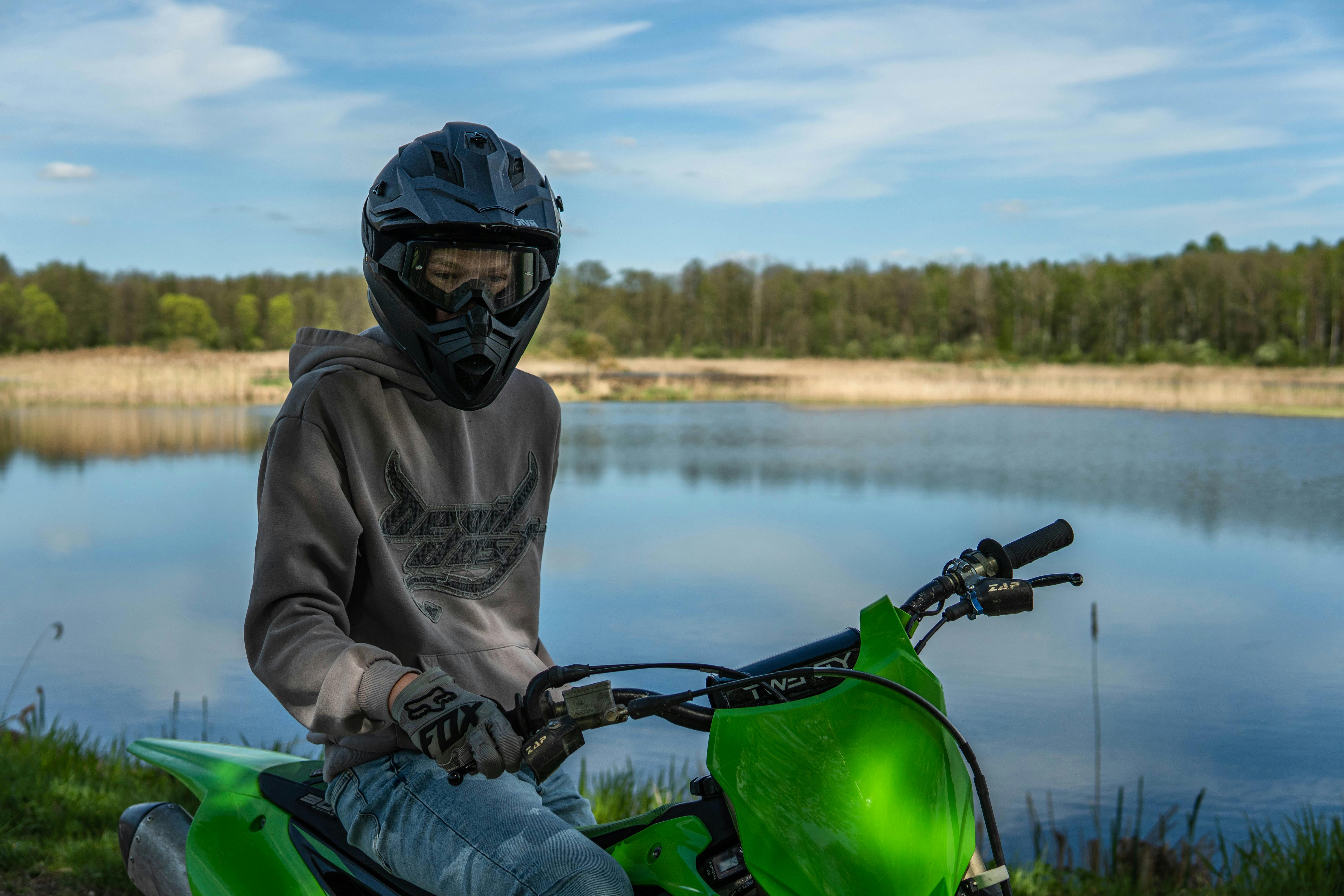 A teenager on a green dirt bike wearing a helmet by a tranquil lake under clear skies.