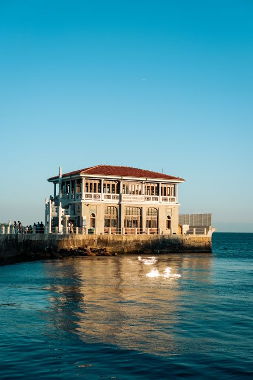Free Elegant seaside building reflecting in calm waters under a clear blue sky. Stock Photo
