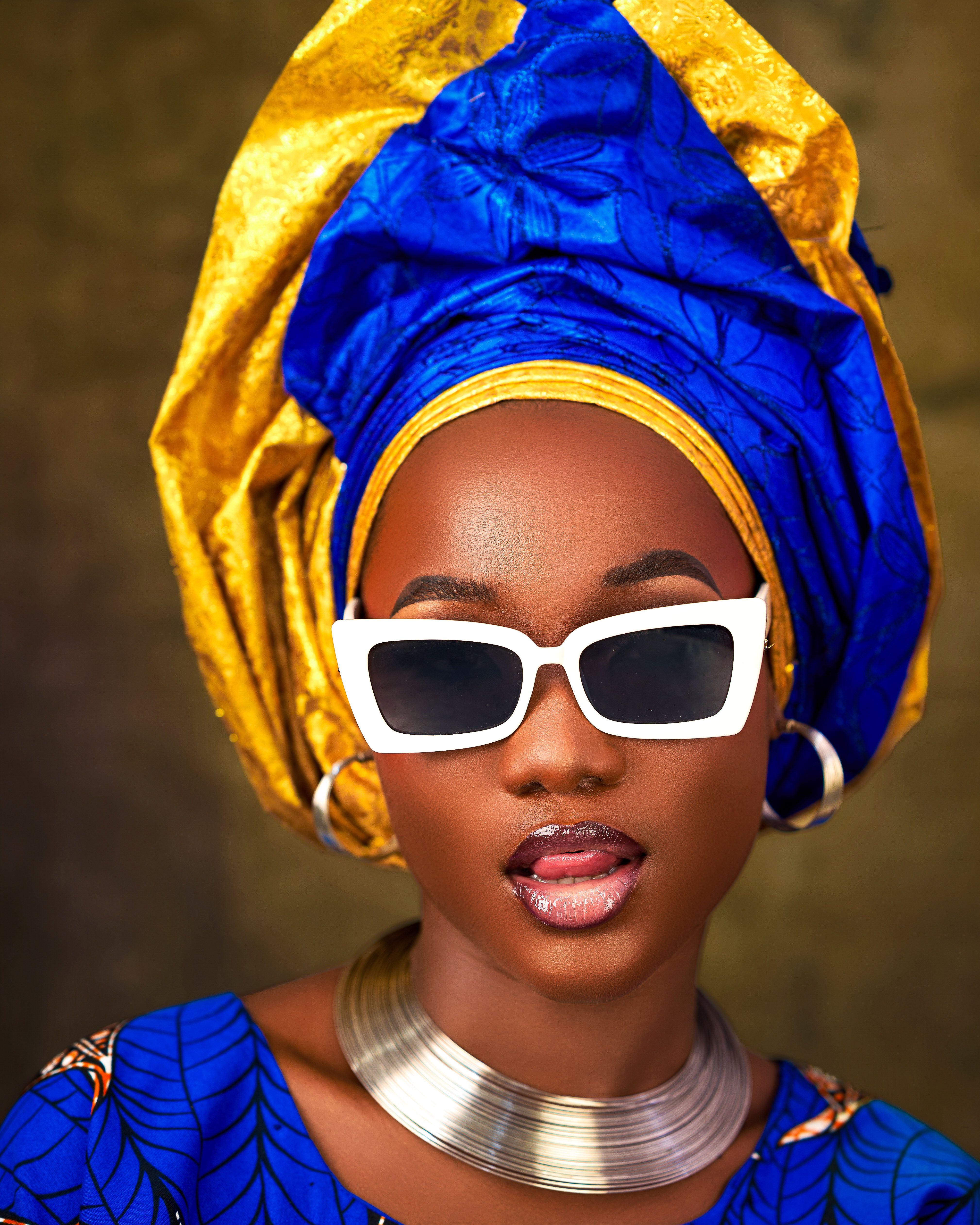 Stylish African woman in blue and gold head wrap wearing white sunglasses and jewelry.