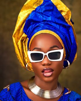 Stylish African woman in blue and gold head wrap wearing white sunglasses and jewelry.