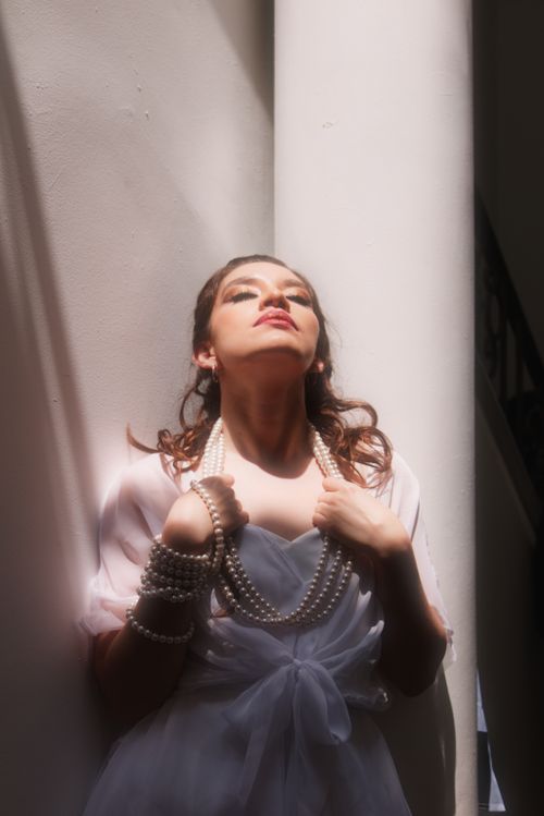 Free A woman in an elegant dress and pearls poses dramatically in sunlight indoors. Stock Photo