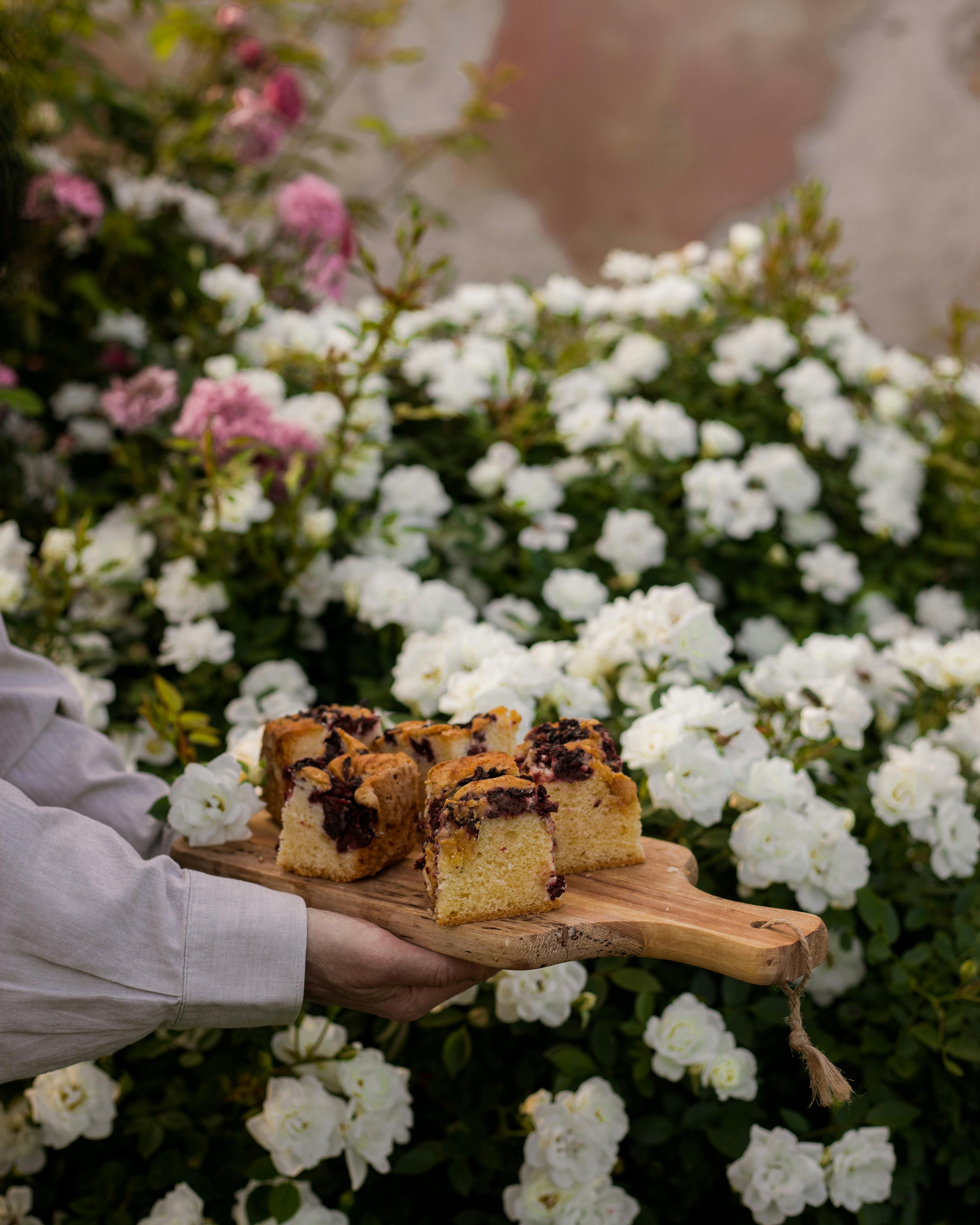 Homemade cake slices on a wooden board amidst vibrant roses in a garden.