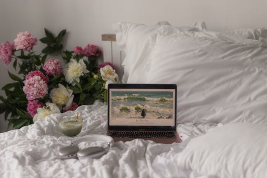 A peaceful bedroom scene with peonies, a laptop, matcha, and headphones for a relaxing atmosphere.
