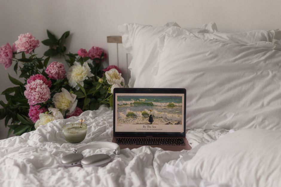 A peaceful bedroom scene with peonies, a laptop, matcha, and headphones for a relaxing atmosphere.