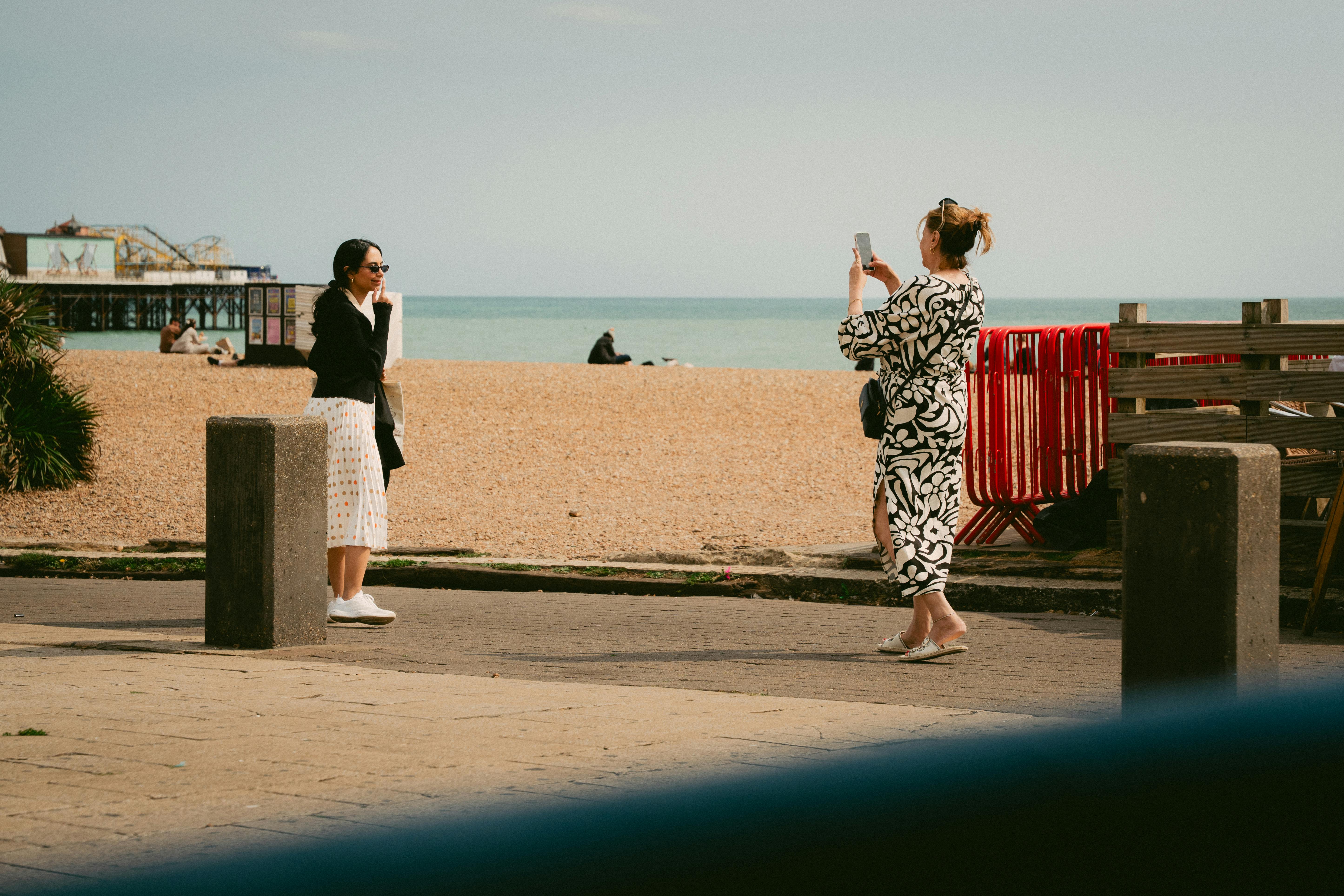 Two women enjoying a sunny day at Brighton Beach, capturing memories with phones.
