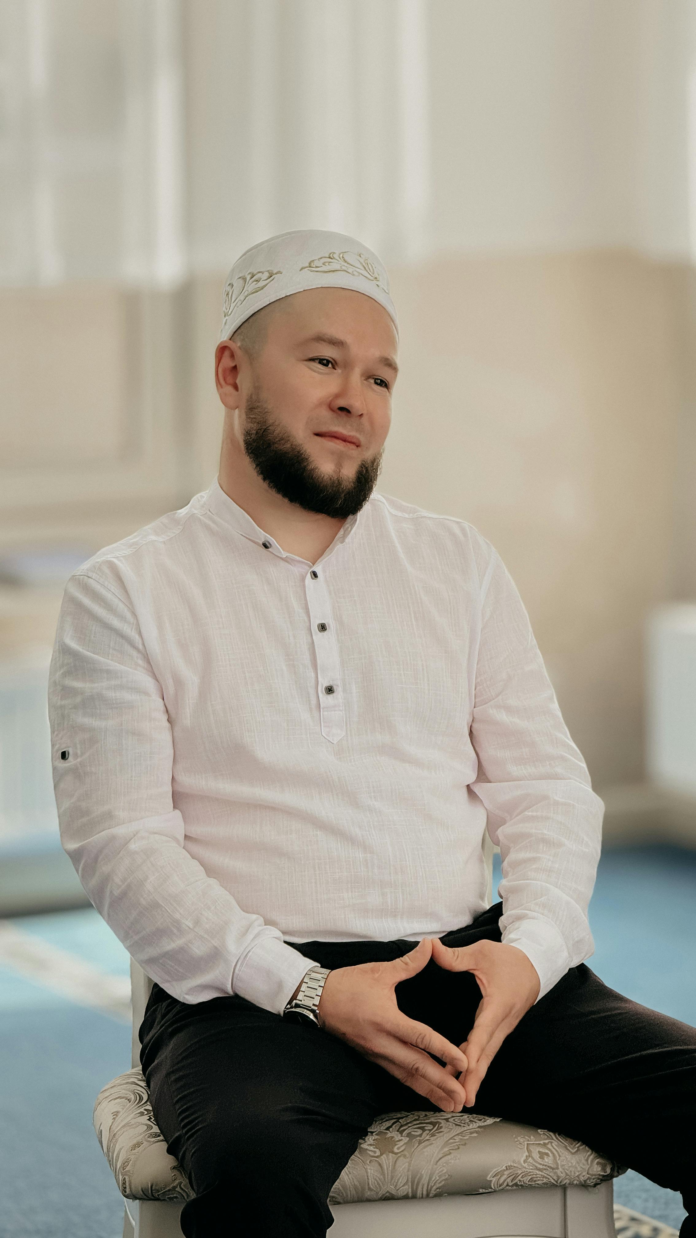 Man sitting indoors, wearing a white shirt and cap, calm expression.