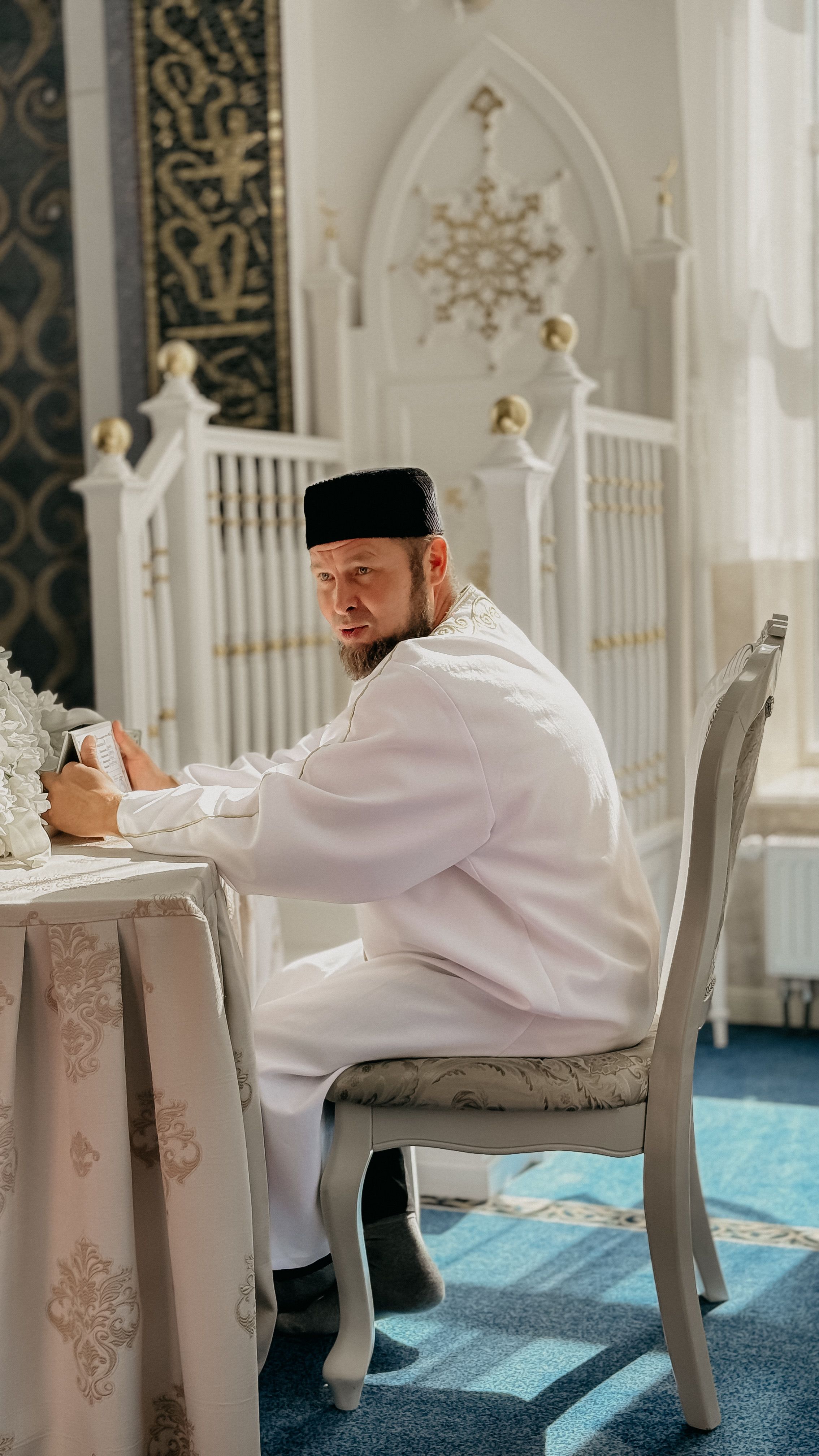 Adult male in traditional dress inside an ornate mosque, reading with sunlight streaming in.