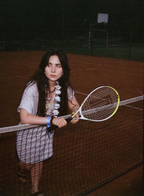 Free Young woman holds a tennis racket on an outdoor clay court at night, exuding a casual and relaxed vibe. Stock Photo