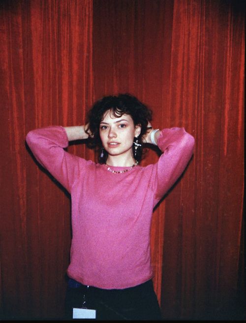 Free Portrait of a young woman in a pink sweater posing stylishly indoors against red velvet curtains. Stock Photo