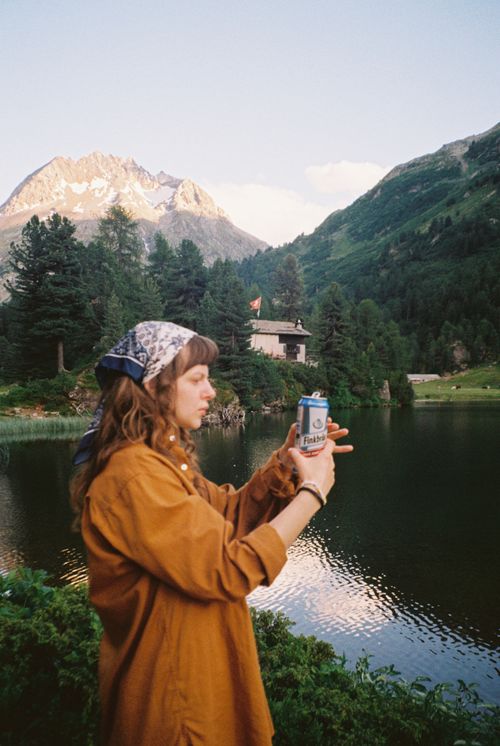 Free A woman holding a can of beer by a lake with mountains at dusk, capturing serenity and leisure. Stock Photo