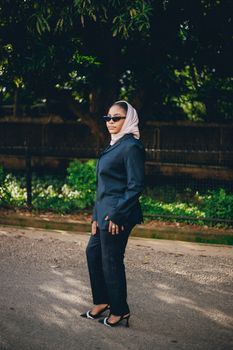 Stylish woman in sunglasses and hijab, standing confidently in a suit outdoors.