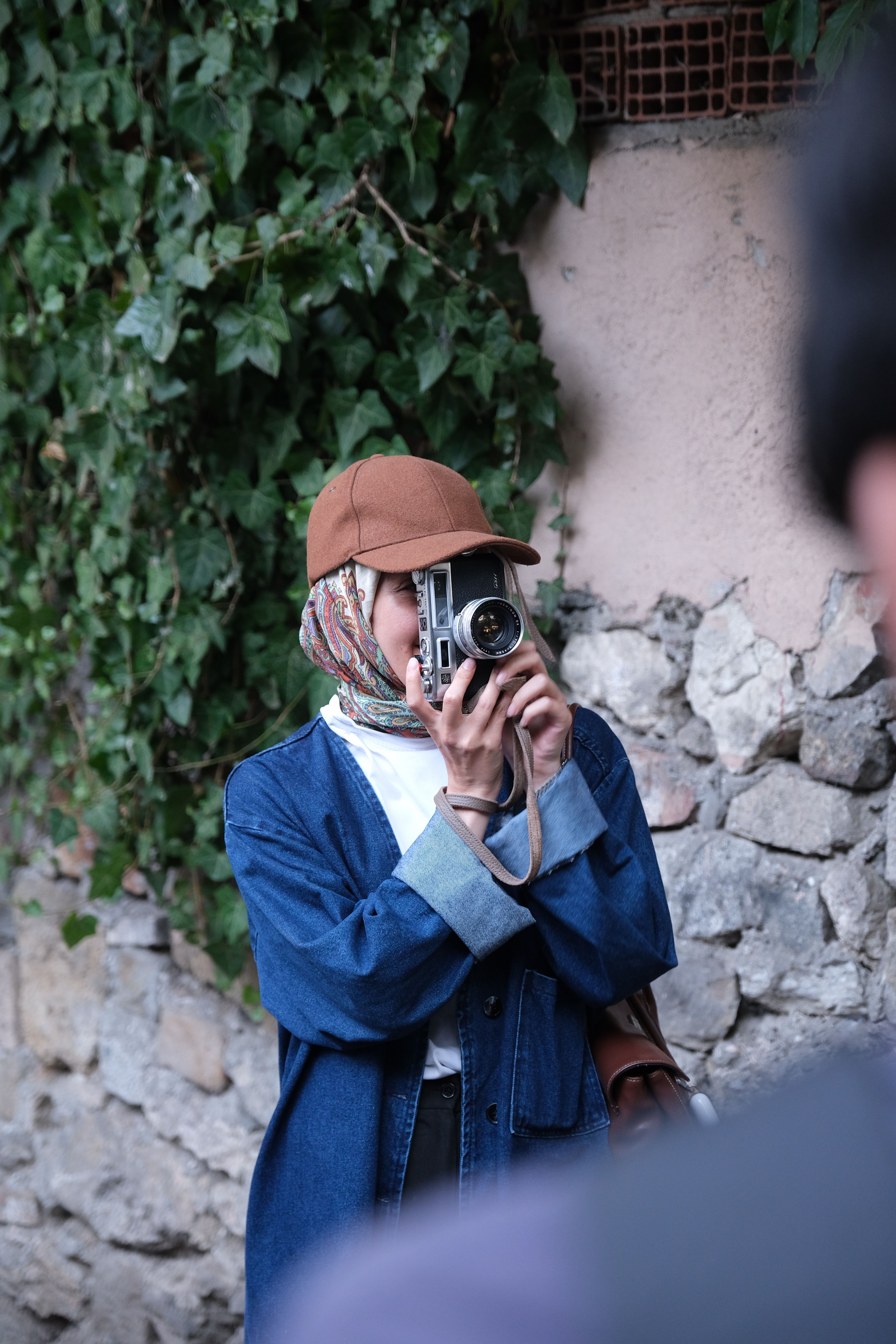 Young woman in denim capturing photos outdoors against stone wall and ivy.