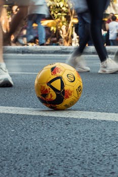 A yellow soccer ball rests on an urban street with blurred pedestrians passing by.
