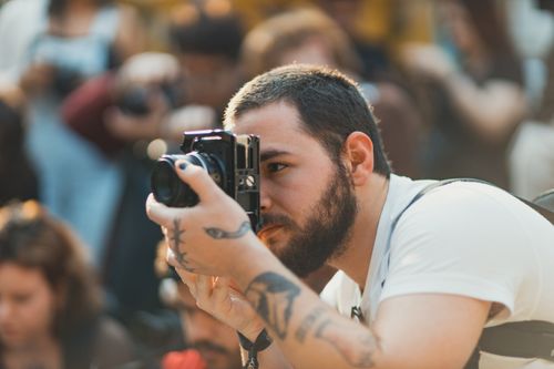 Free Man with tattoos focuses intently on capturing a moment with a camera at a busy outdoor event. Stock Photo