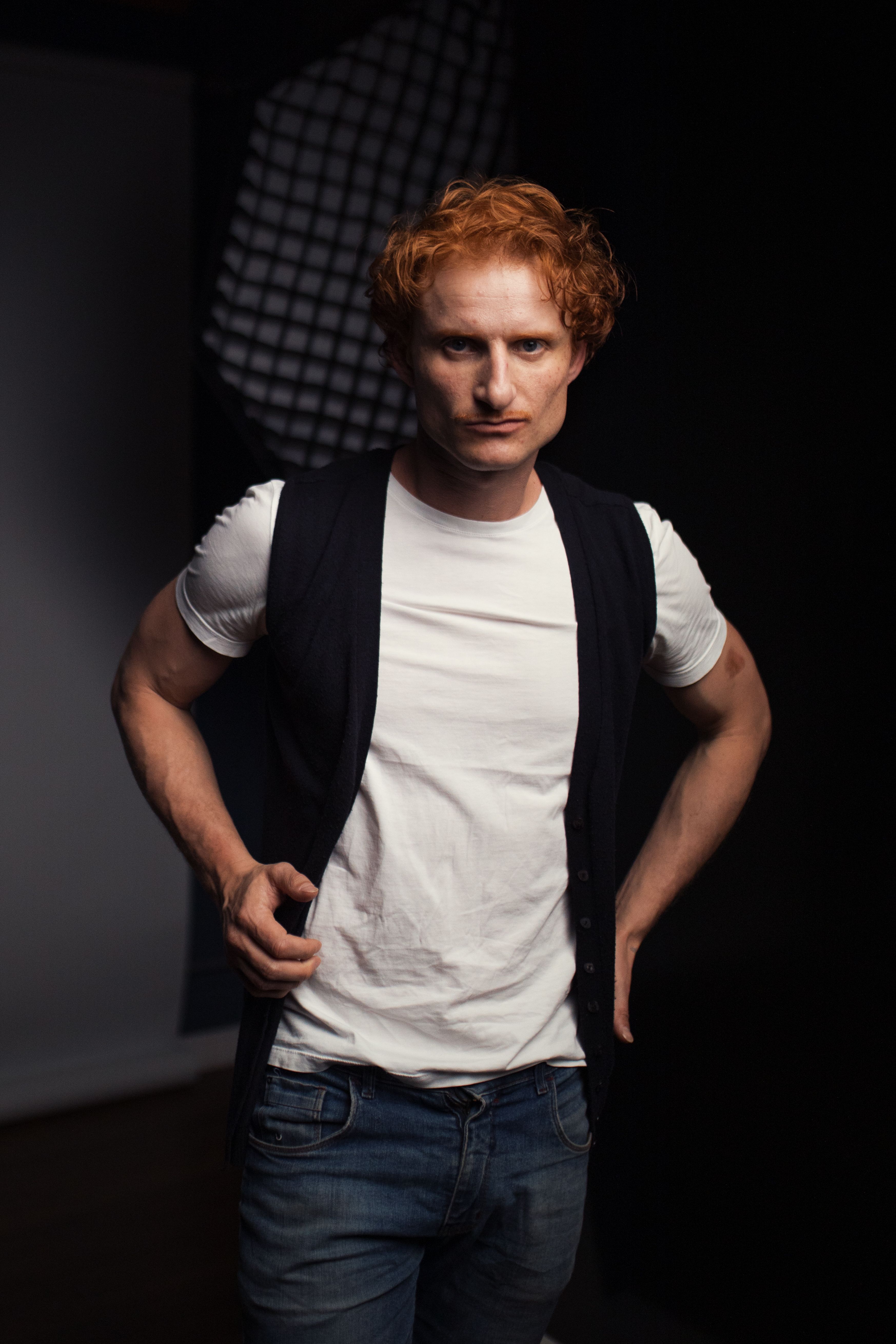 Young man with red hair poses confidently in a studio with soft lighting creating a dramatic effect.