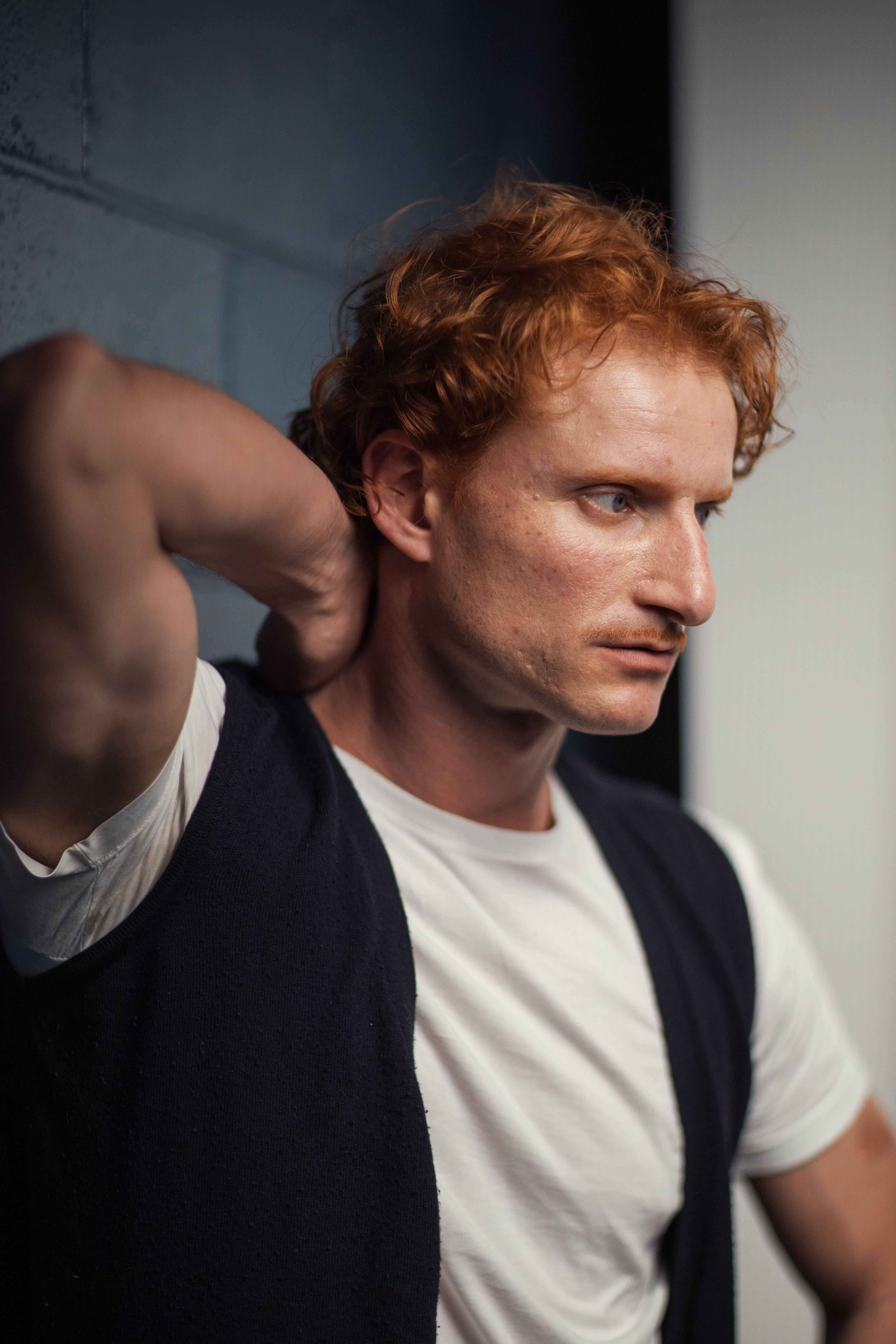 Red-haired man in casual attire poses against a wall, deep in thought.