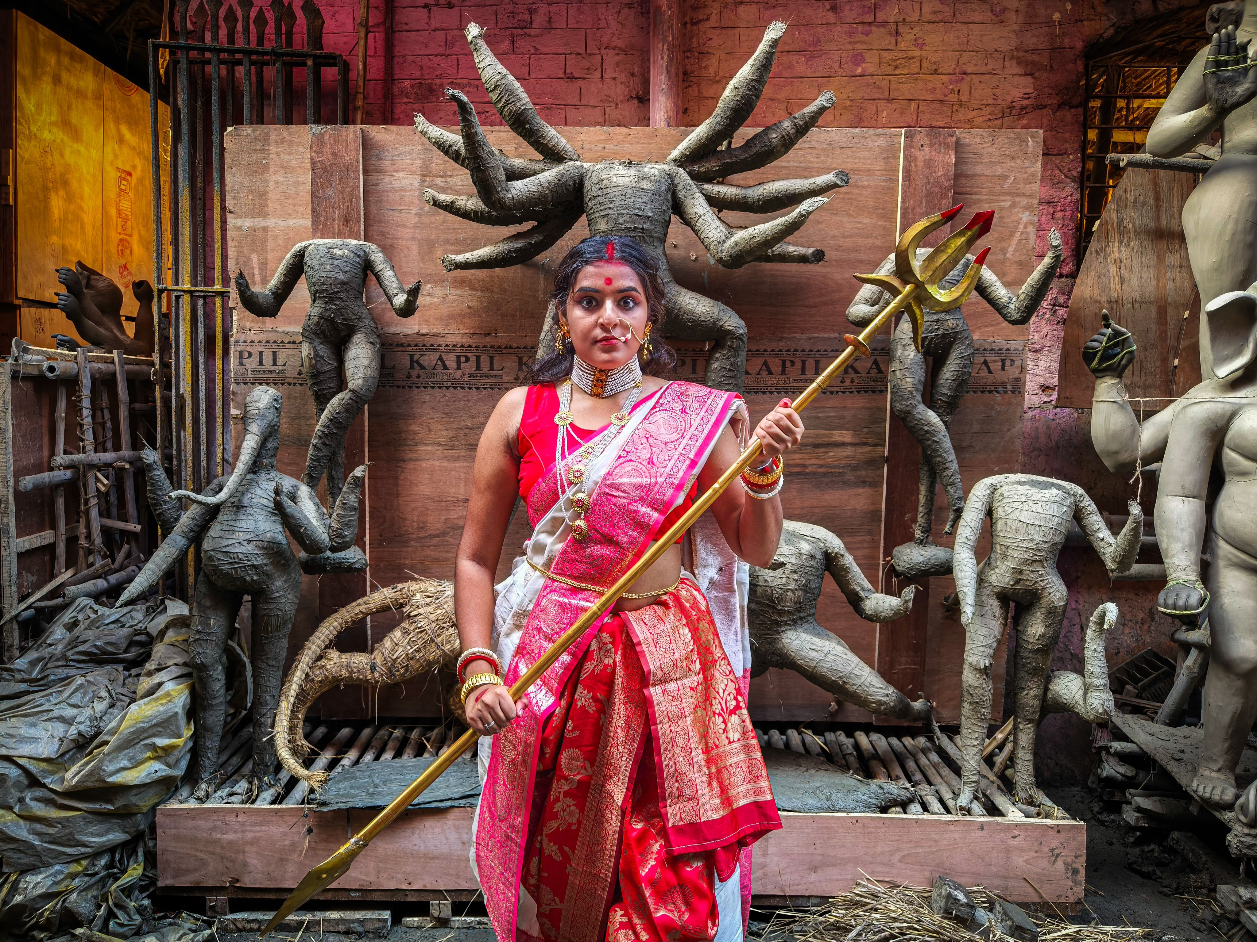A woman in traditional attire poses with a trident amidst clay idols in a Kolkata workshop during Durga Puja preparations.