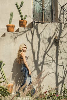 Woman standing in a sunlit courtyard with cacti, showcasing fashion and shadow play.