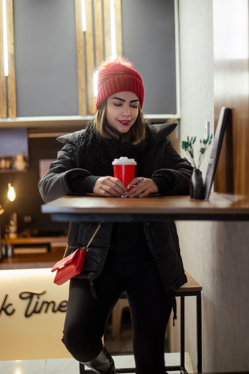 Stylish woman in red beanie enjoying coffee indoors, creating a cozy vibe.