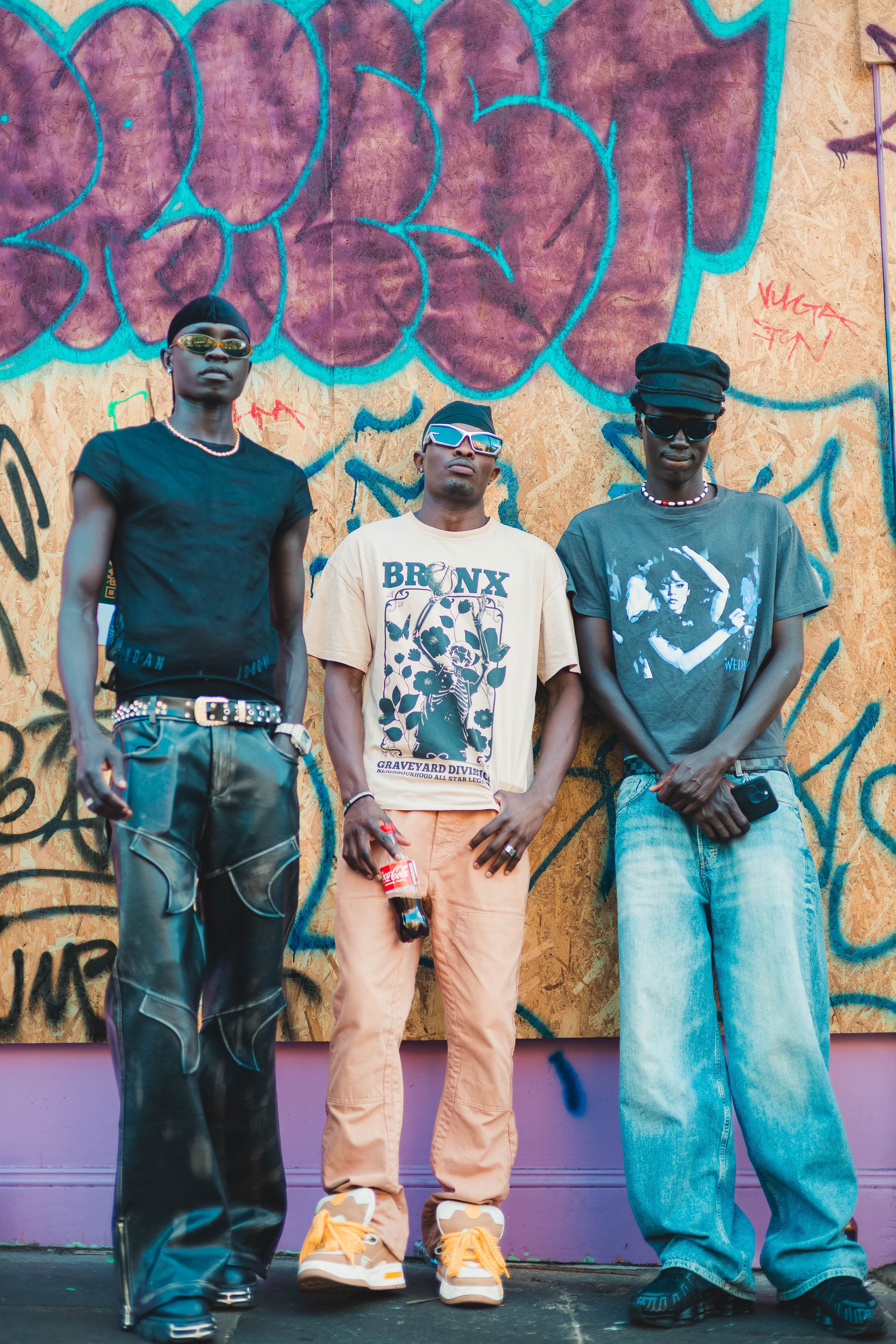 Three men pose in urban streetwear against a graffiti wall in London.