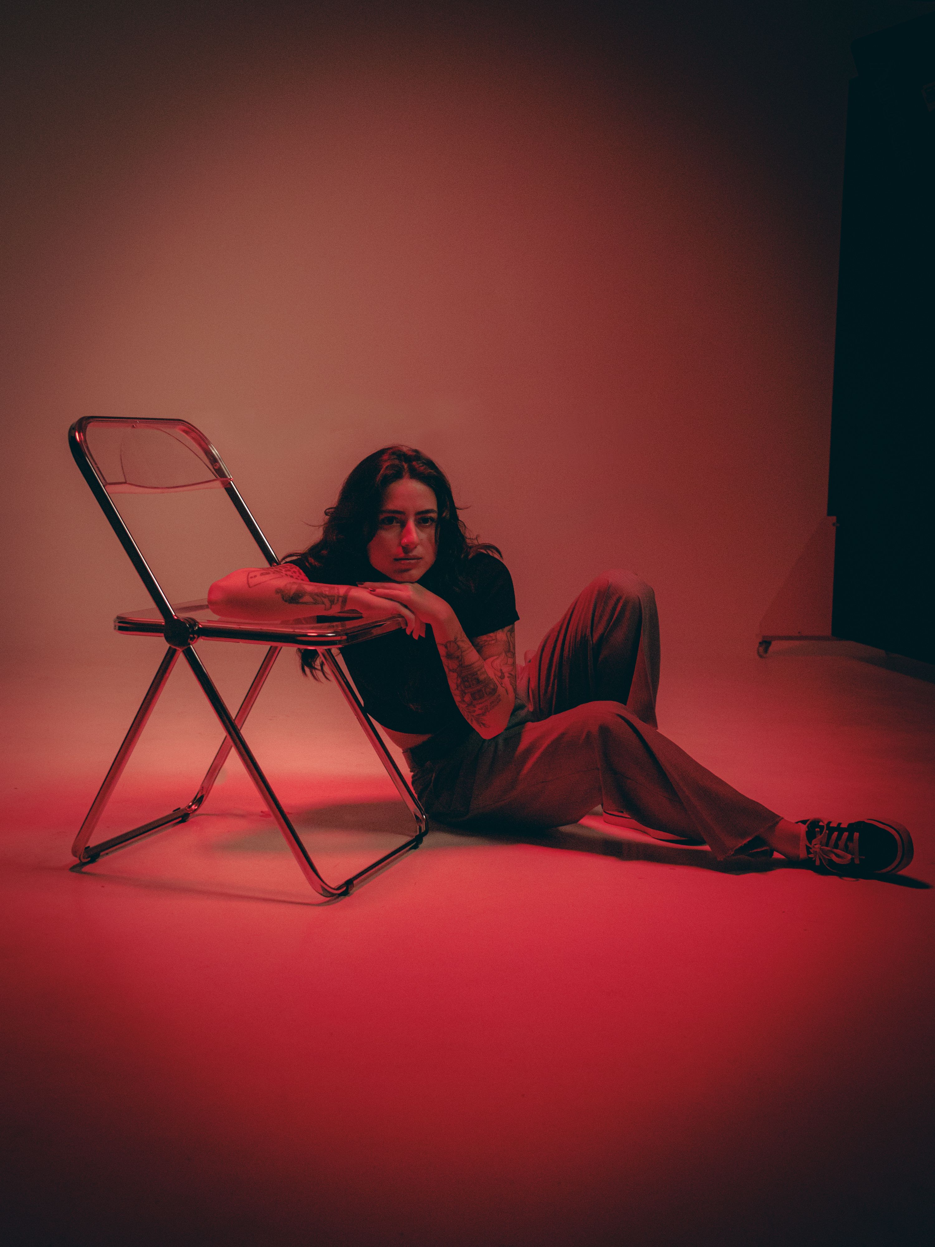 A person seated on the floor with red lighting, leaning on a chair in a studio setting.