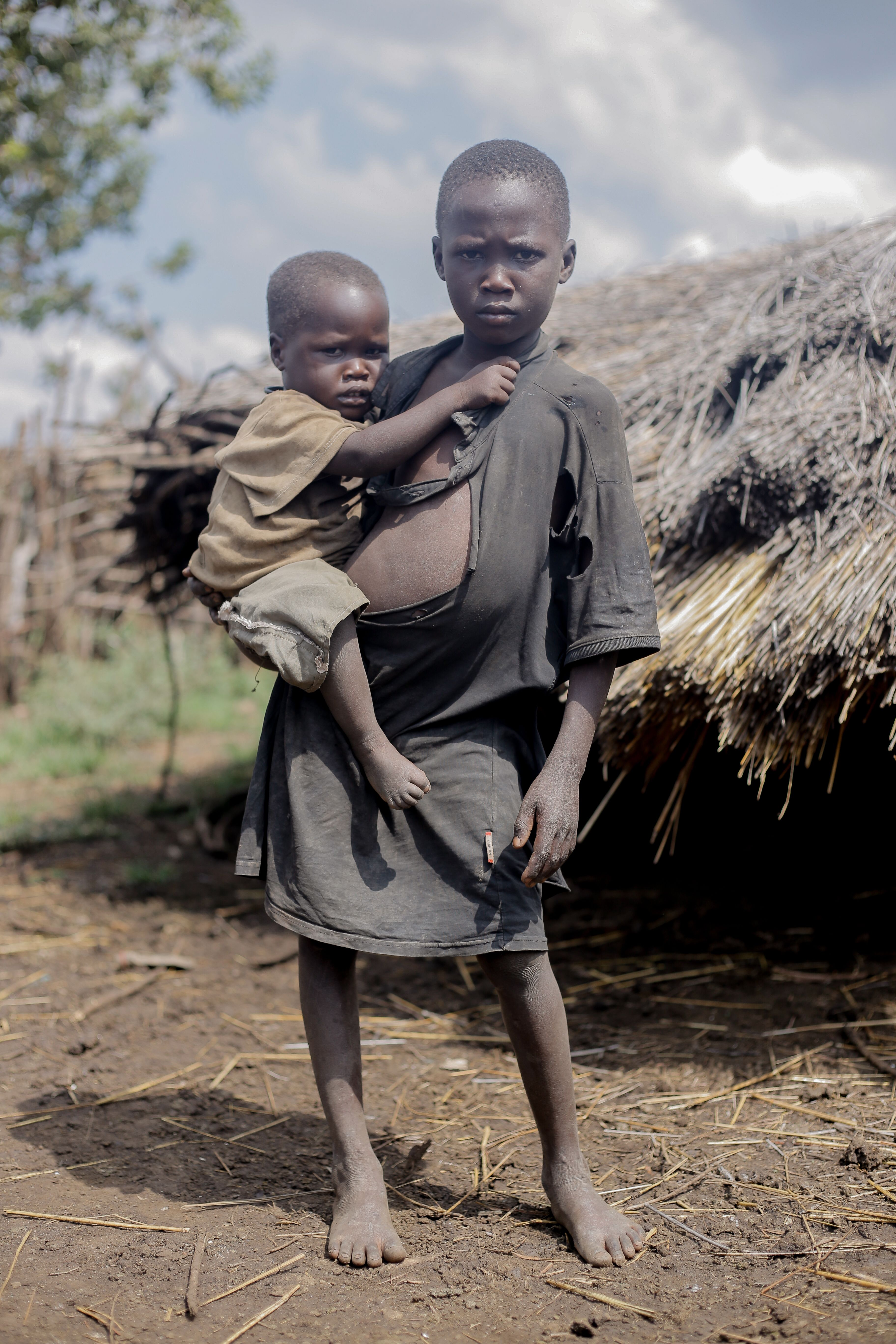 Two young African siblings in a rural village setting, standing in front of thatched huts.