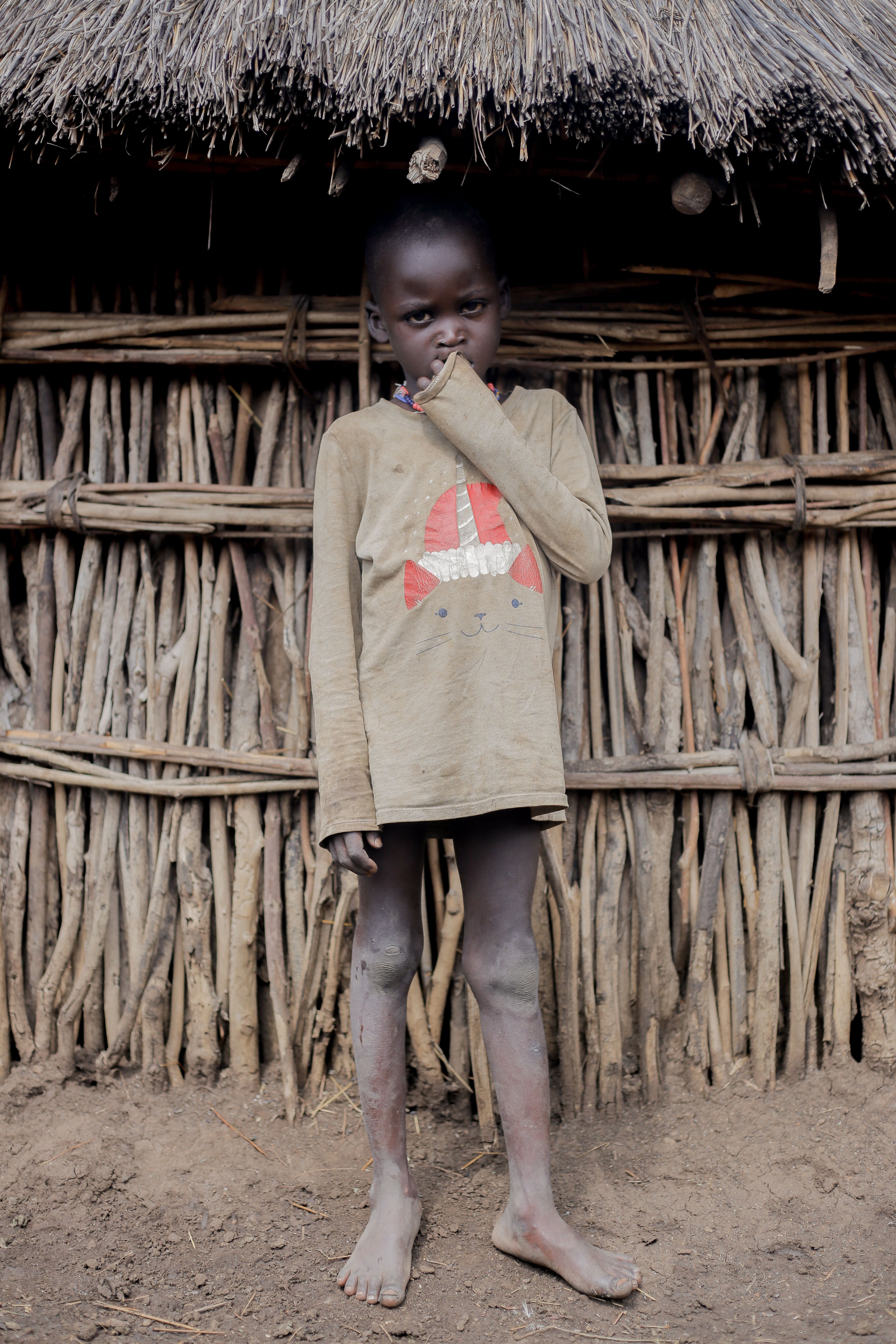 A young child in casual clothing stands barefoot against a rustic wooden wall, conveying simplicity and innocence.